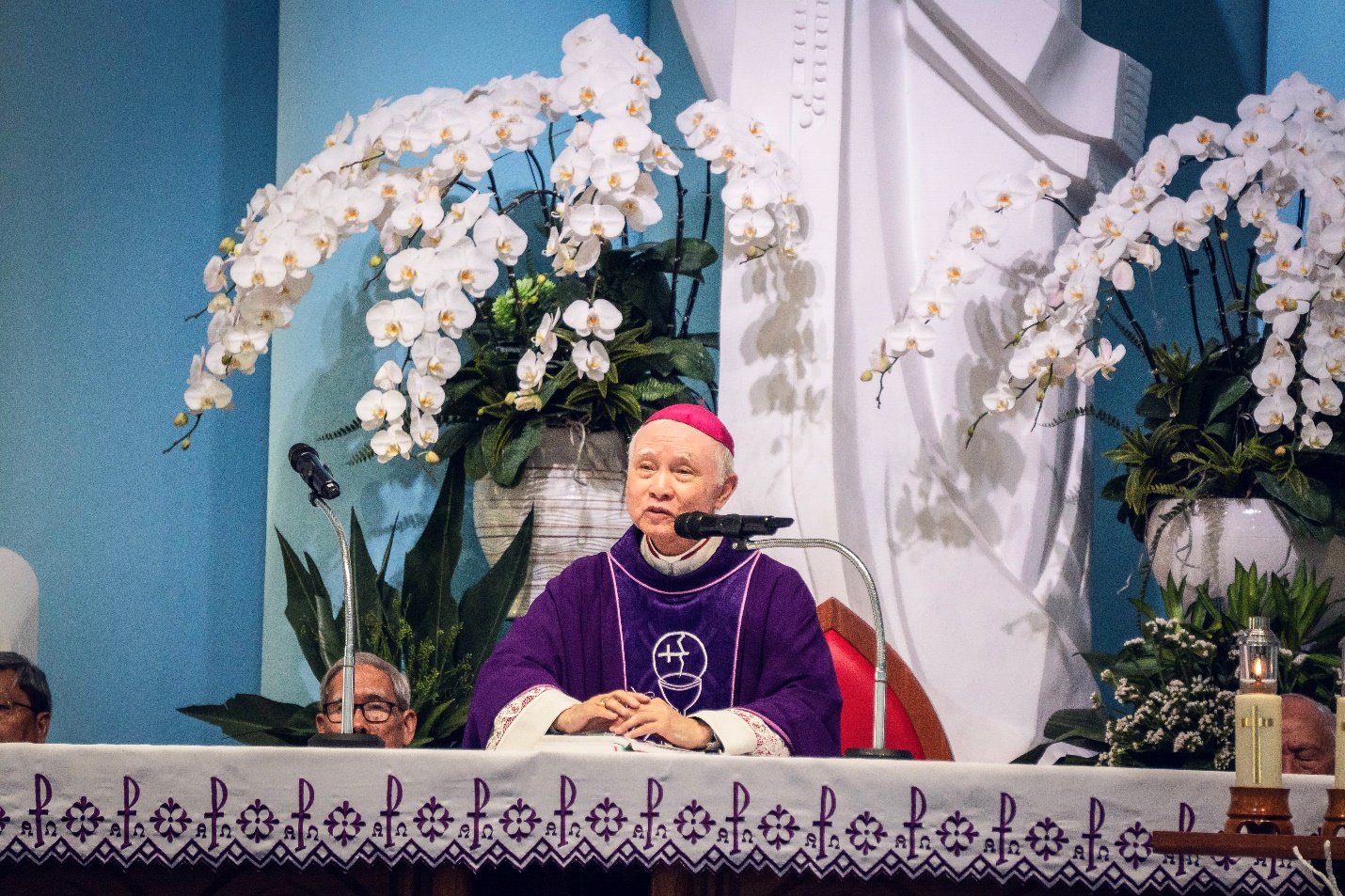 A person in a purple robe sitting at a table with a microphone Description automatically generated